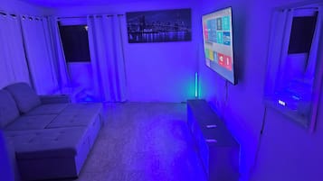 Un salon avec un canapé, un téléviseur fixé au mur et une fenêtre avec des rideaux.