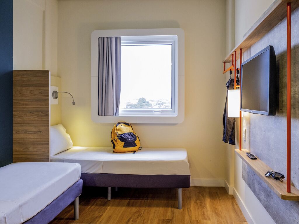 Habitación pequeña con cama, un banco, una ventana con cortinas, un televisor montado en la pared y una mochila sobre el banco.