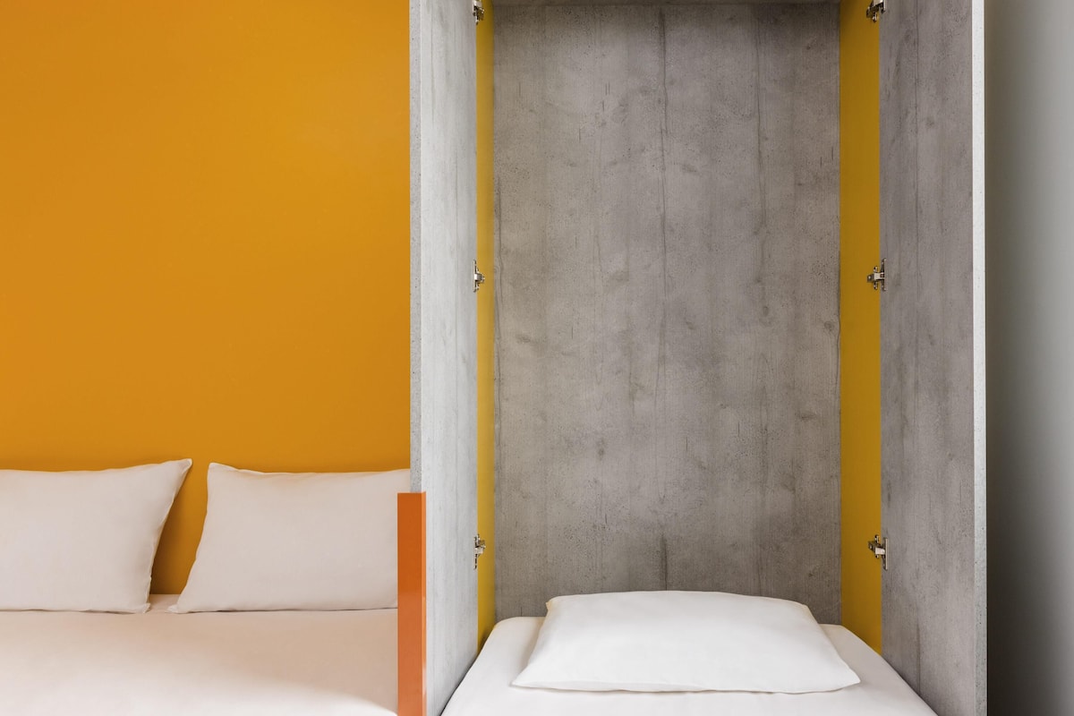 A room with a bed, a yellow wall, and a grey concrete partition.