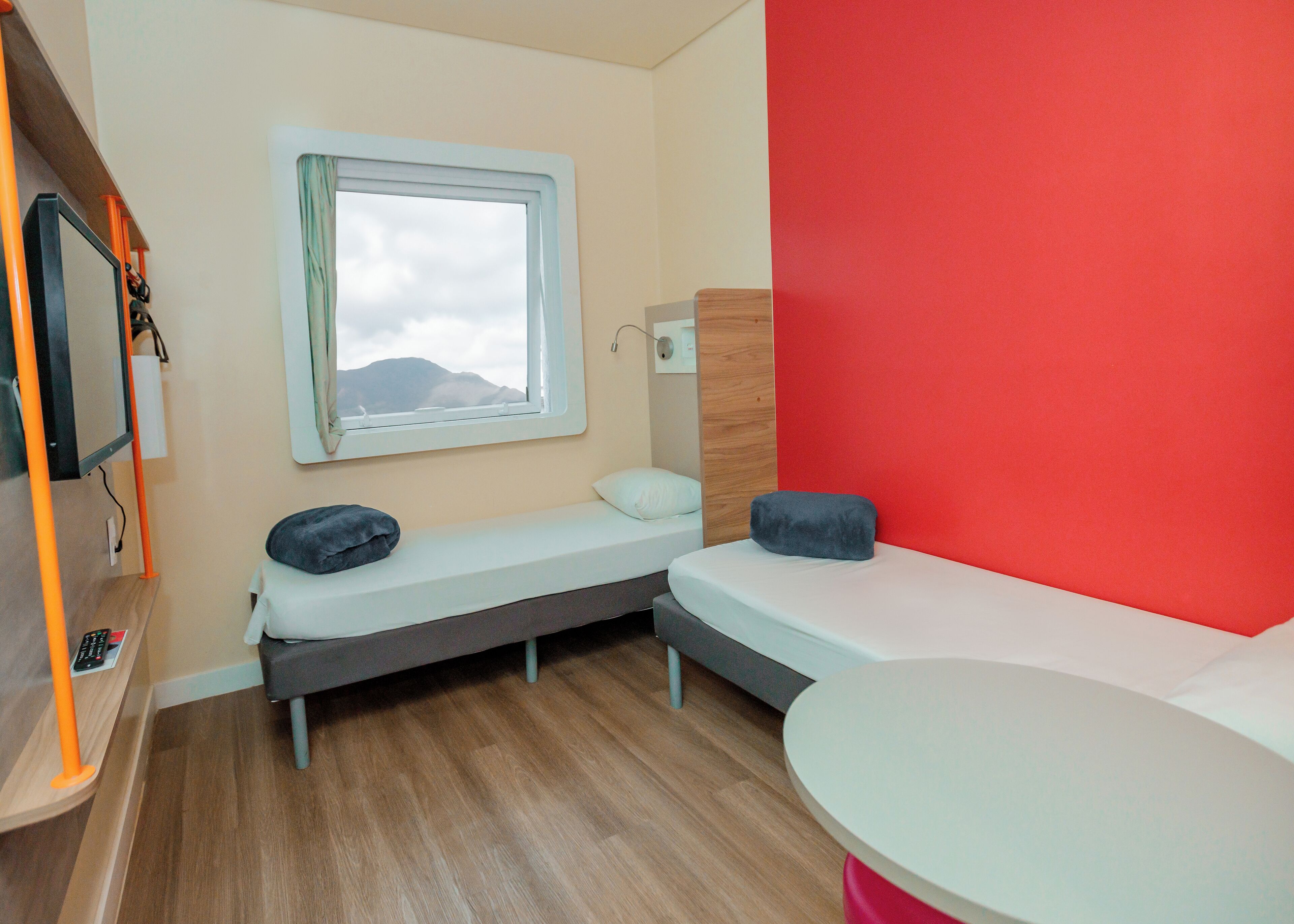 Habitación compacta con dos camas, una mesita y ventana con vistas a montañas.