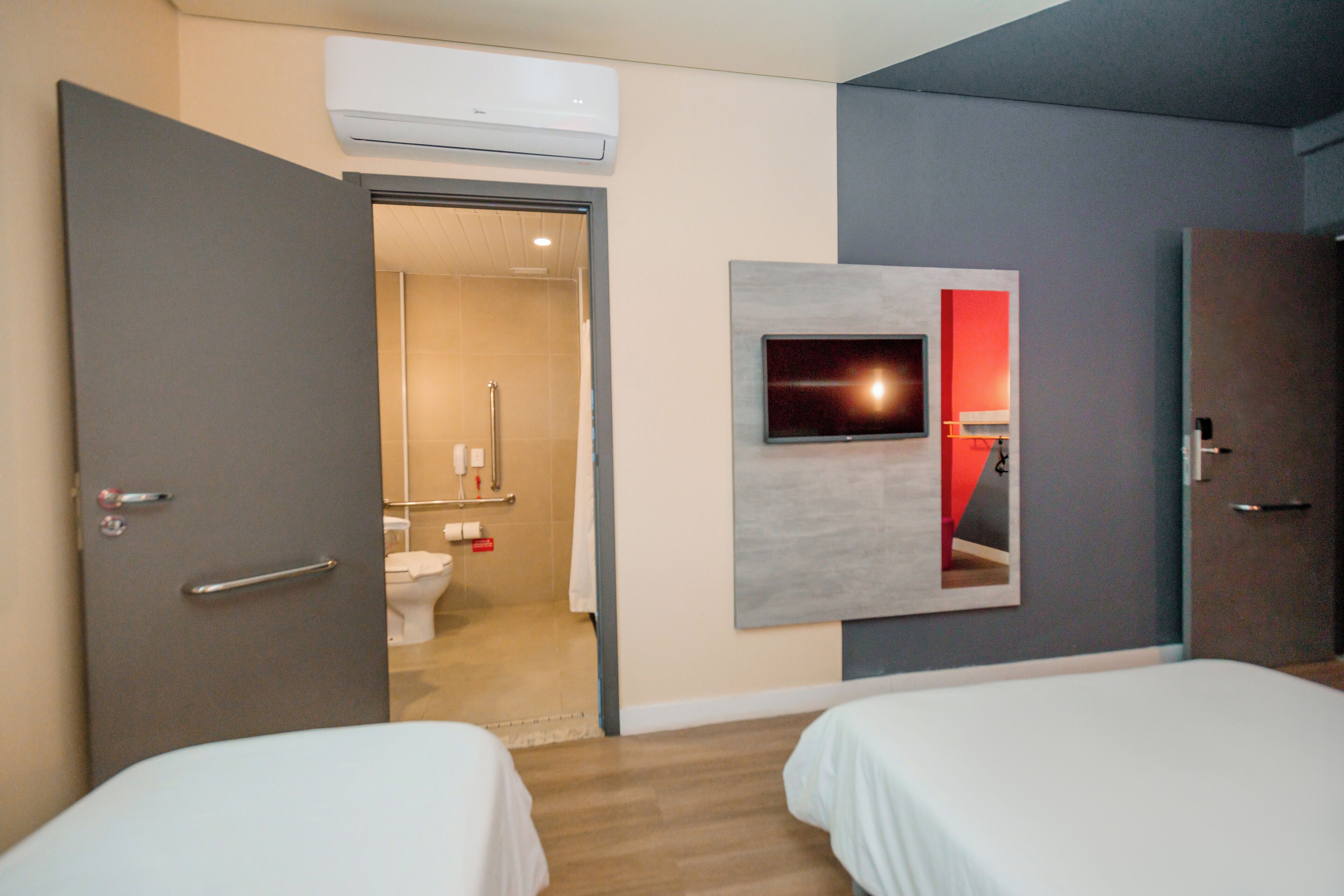 Una habitación de hotel con dos camas, un televisor montado en la pared, un equipo de aire acondicionado sobre la puerta y un baño visible a través de una puerta abierta.