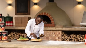 A chef preparing pizza in front of a brick oven with a sign above reading "DE LUCAS".