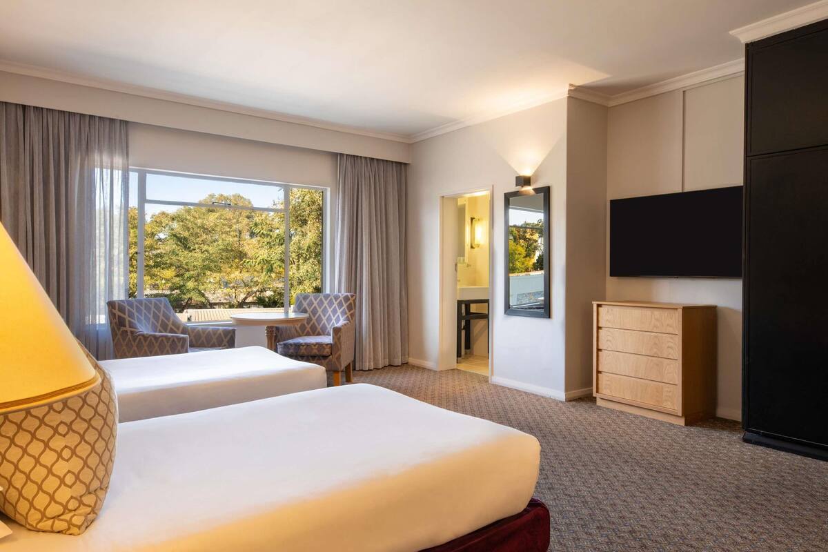 A hotel room with two beds, a TV, a desk, and a view of trees outside.