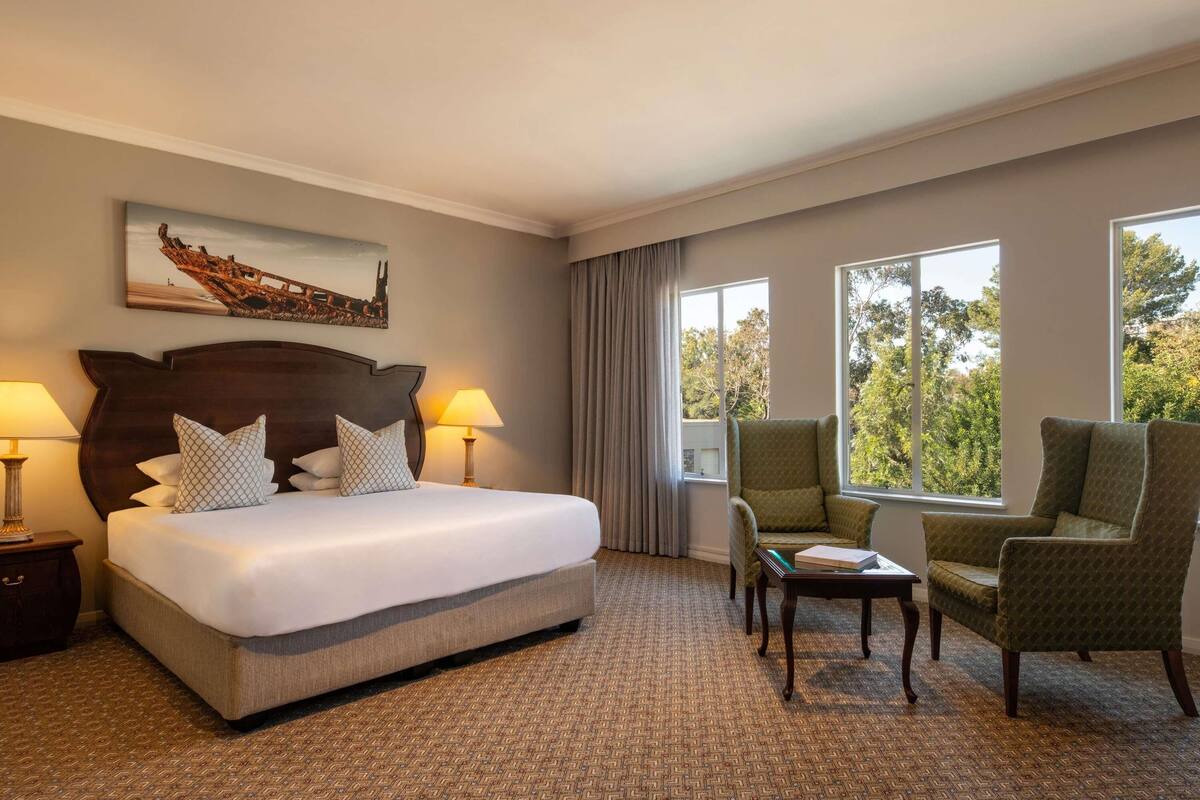 A hotel room with a large bed, two bedside lamps, a small table, two armchairs, and a window with a view of trees.
