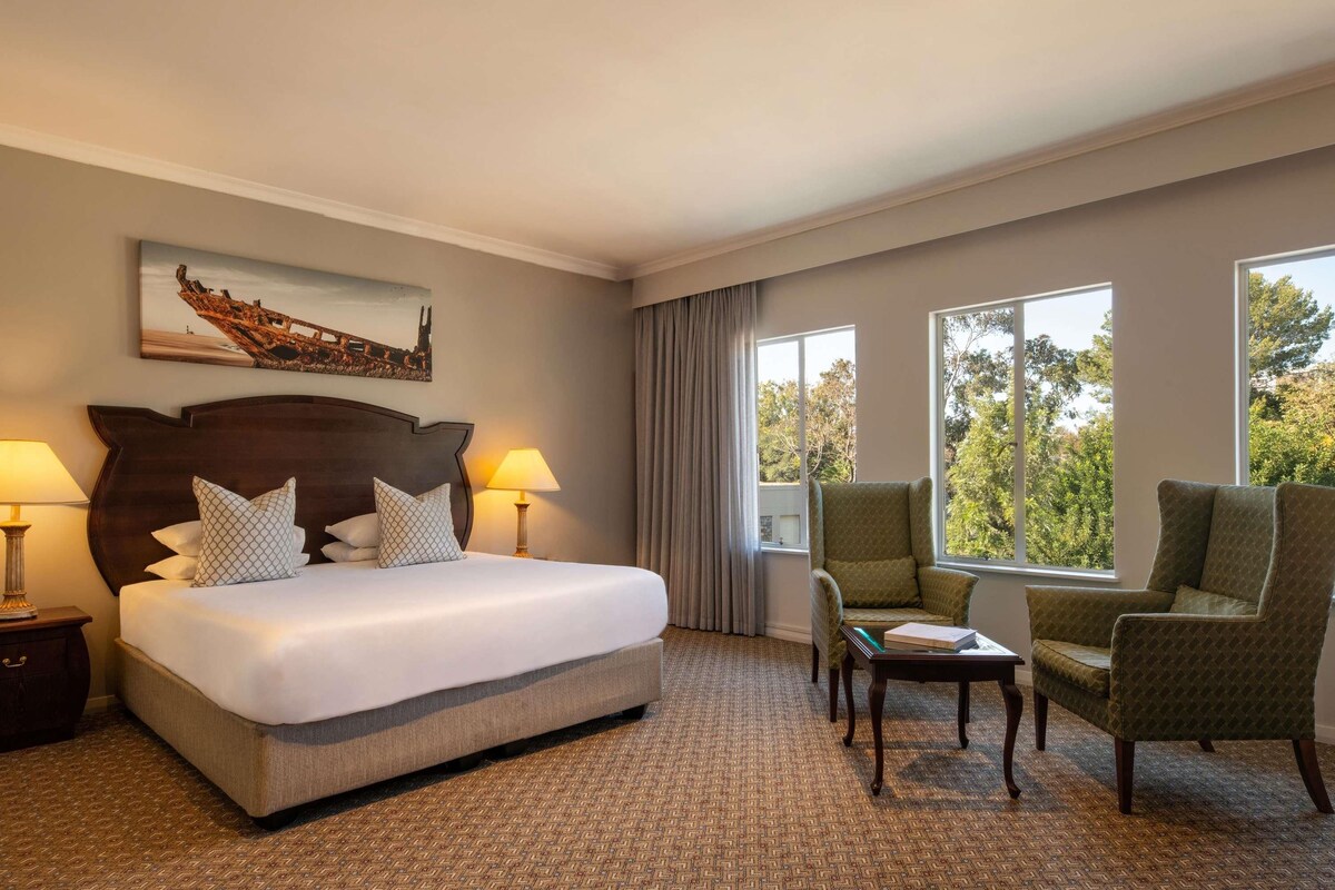 A hotel room with a large bed, two bedside lamps, a small table, two armchairs, and a window with a view of trees.