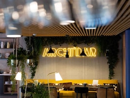Interior de restaurante con letrero de neón, asientos, iluminación colgante y plantas.