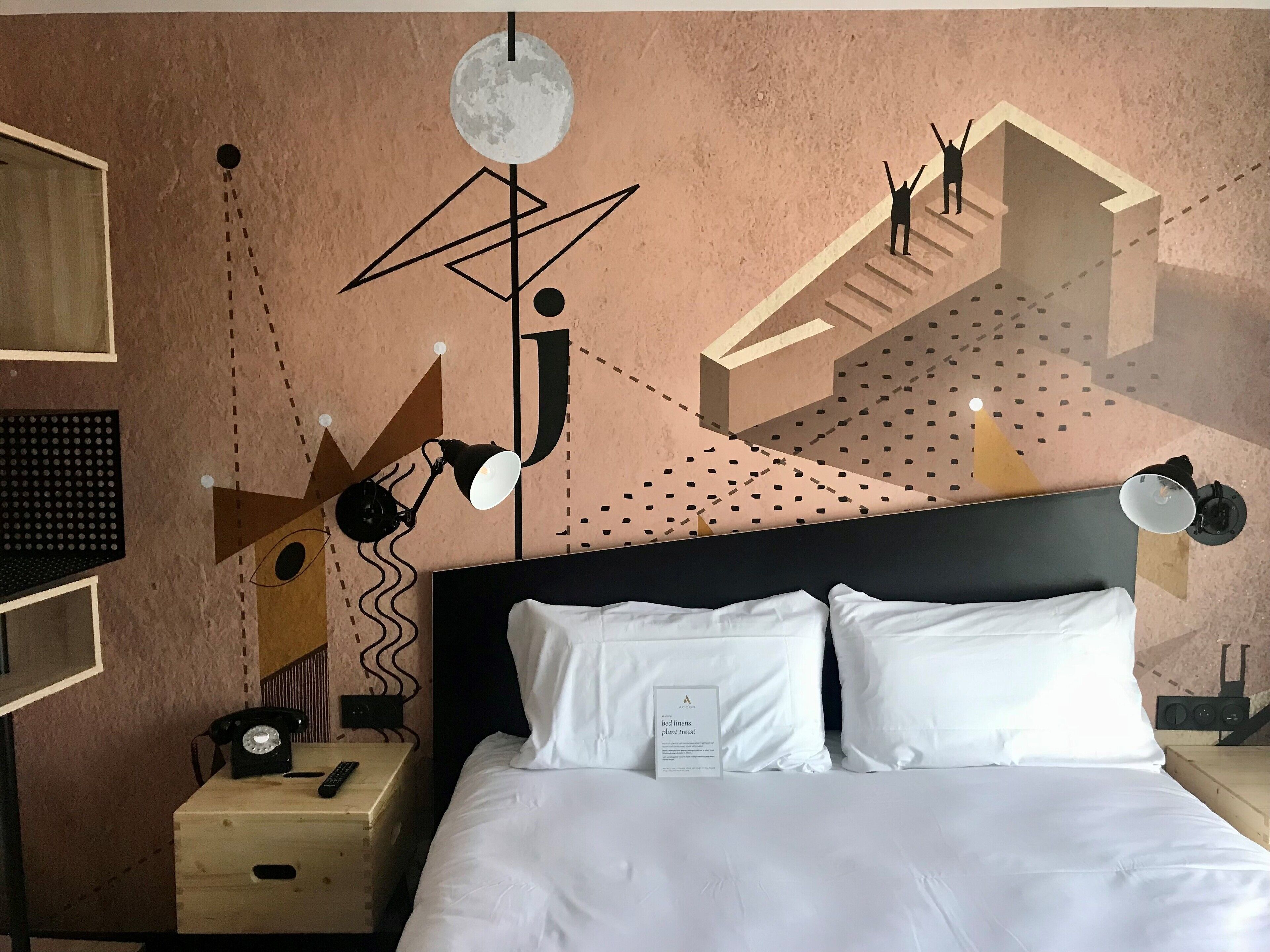 Hotelkamer met een bed, nachtkastje met telefoon en afstandsbediening, wand met abstracte kunst en een plafondlamp.