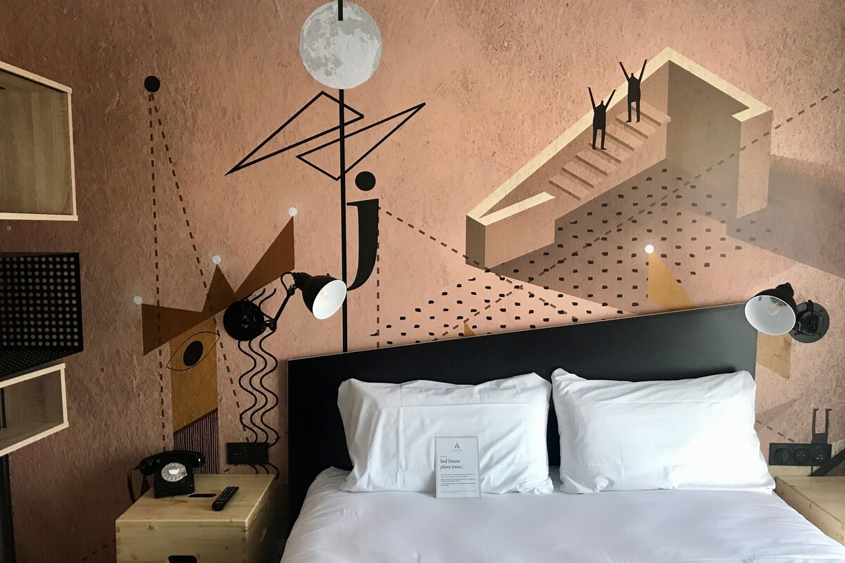 Una habitación de hotel con una cama, una mesita de noche con teléfono y control remoto, una pared con arte abstracto, e iluminación en el techo.