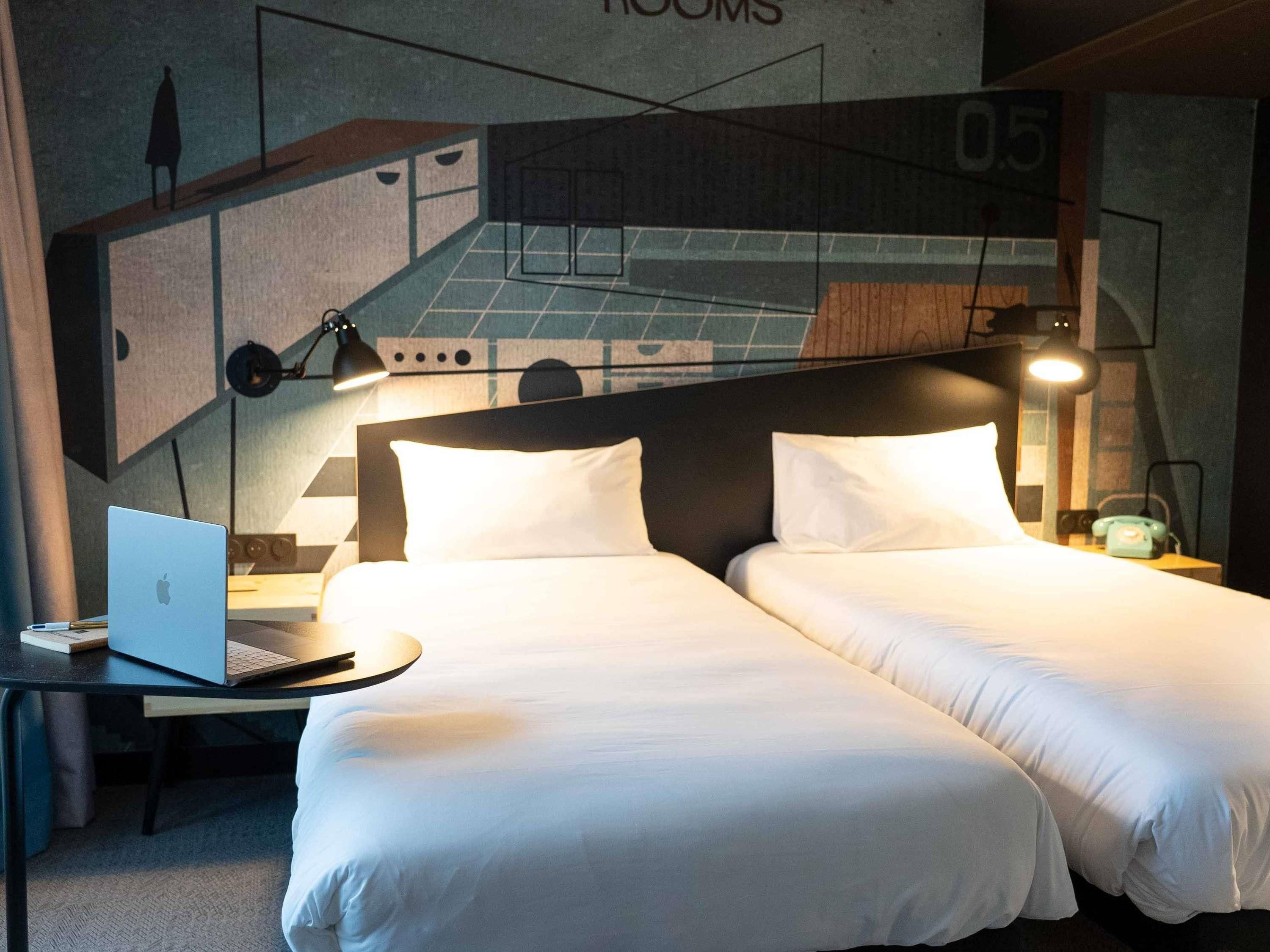 A hotel room with two beds, a desk with a laptop, and a wall with a geometric design.