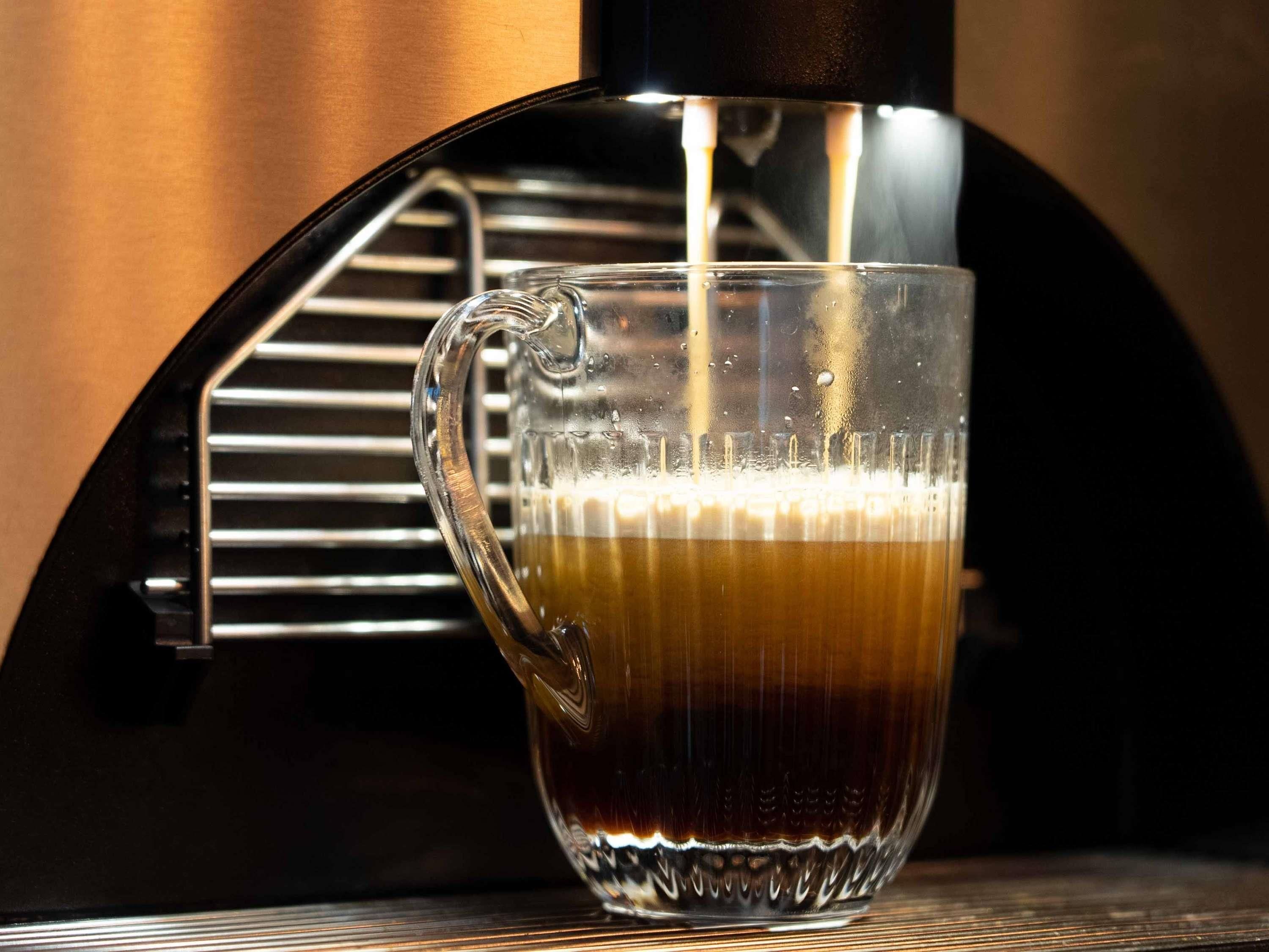 A clear glass coffee cup being filled with a dark liquid, likely coffee, from a black coffee machine.