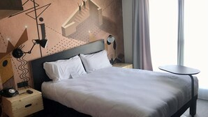 Una habitación de hotel moderna con una cama grande, una mesita de noche con lámpara, una pequeña mesita auxiliar de madera y una mesa redonda junto a la ventana.