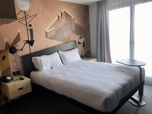 Una habitación de hotel moderna con una cama grande, una mesita de noche con lámpara, una pequeña mesita auxiliar de madera y una mesa redonda junto a la ventana.