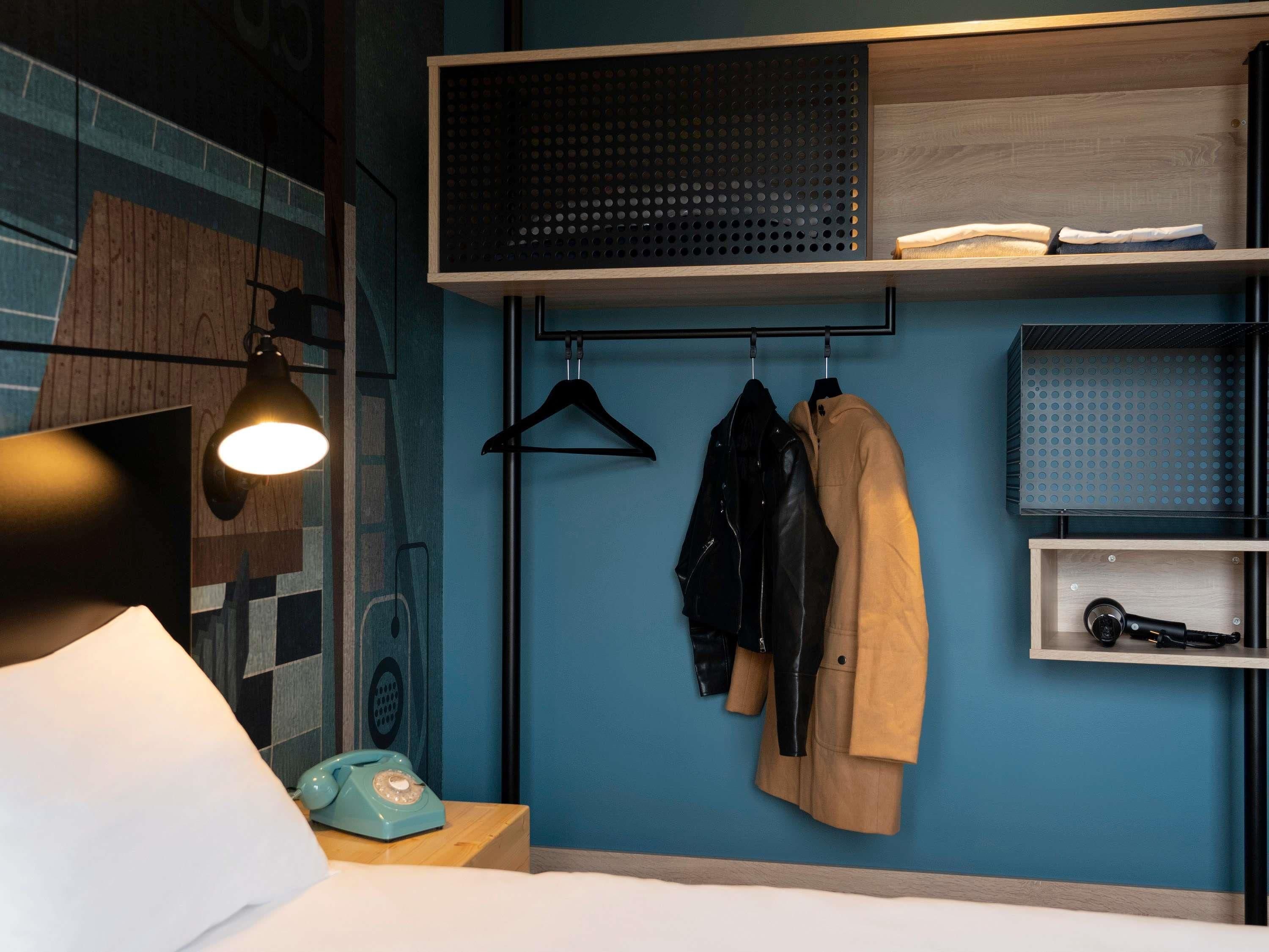 Habitación de hotel con cama, perchero, escritorio con teléfono y estante en la pared con aspiradora.