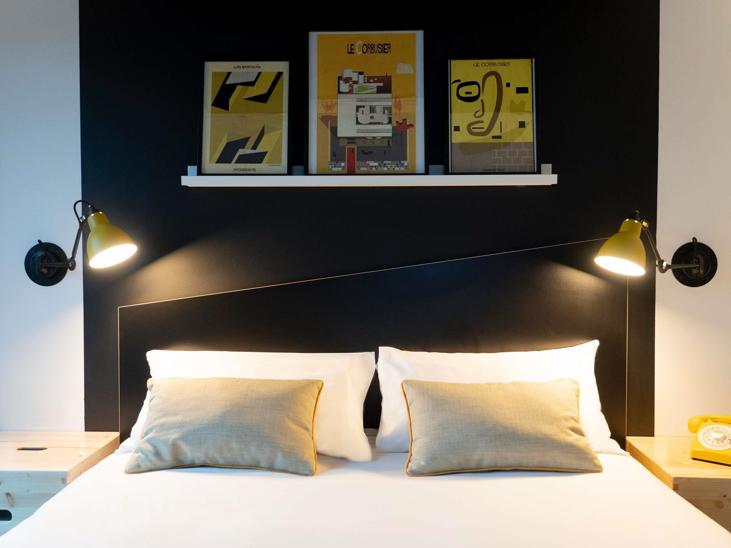 A hotel room with a bed, two bedside lamps, a nightstand with a telephone, and three framed posters on the wall.