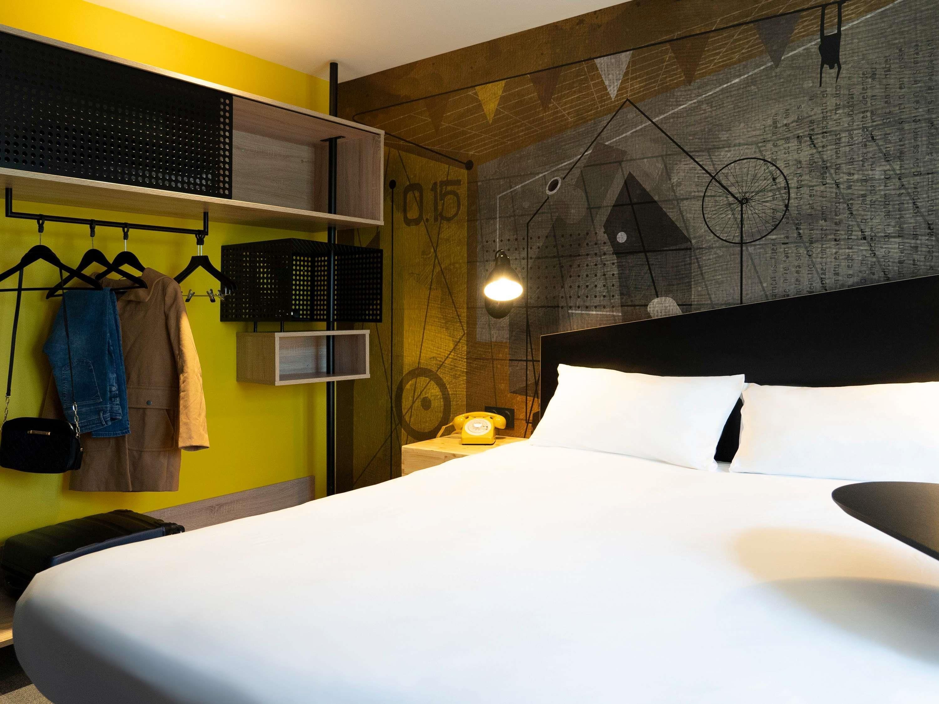 Une chambre d’hôtel moderne dotée d’un grand lit, d’une étagère suspendue pour les vêtements, d’un haut-parleur fixé au mur et d’un décor mural géométrique original.