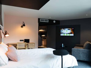 Quarto de hotel moderno com uma cama, escrivaninha, cadeira, TV e armário.