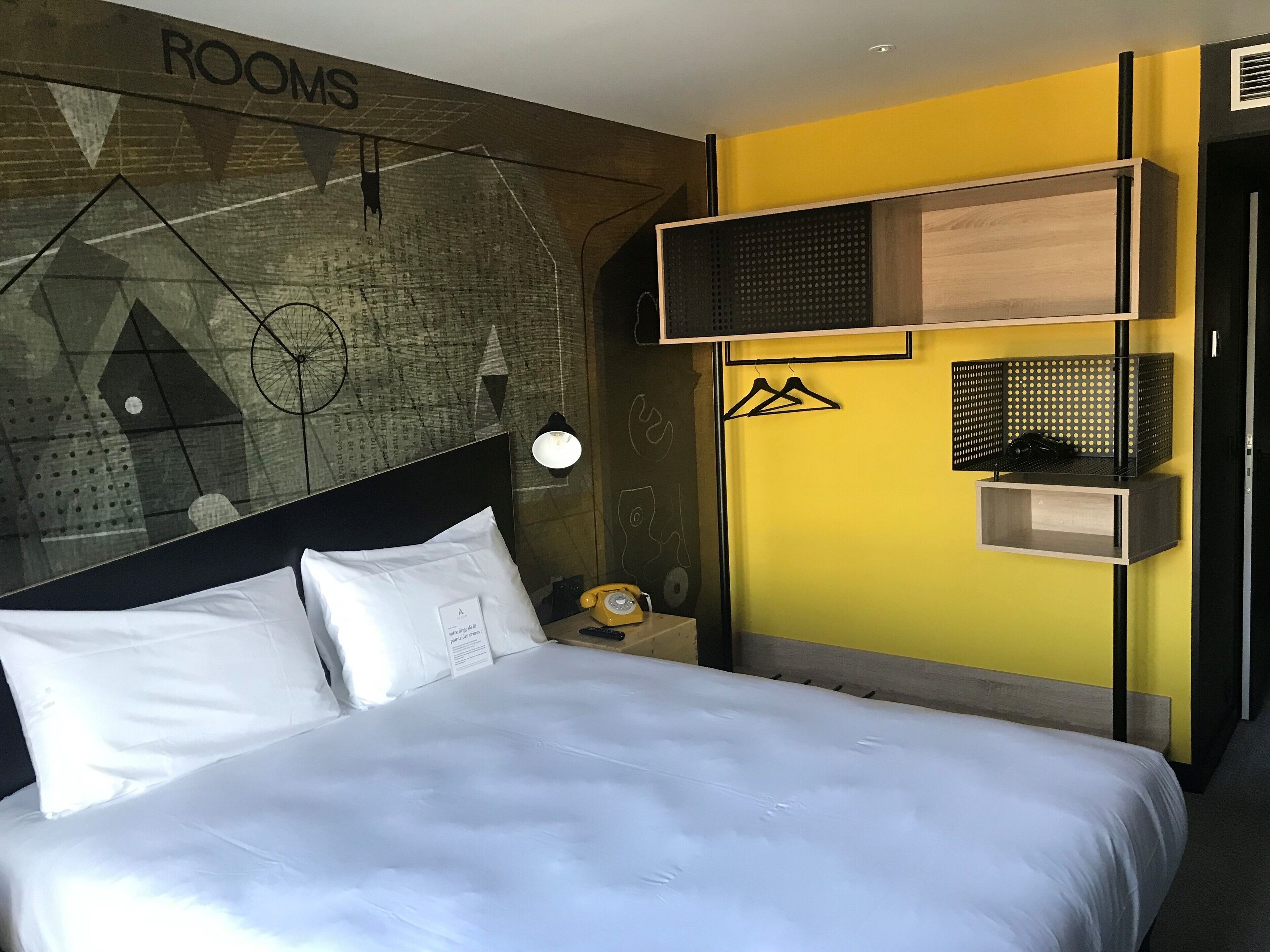 A hotel room with a bed, a wall-mounted TV, a bedside table with a lamp, and a wall with a geometric design and the word "ROOMS".