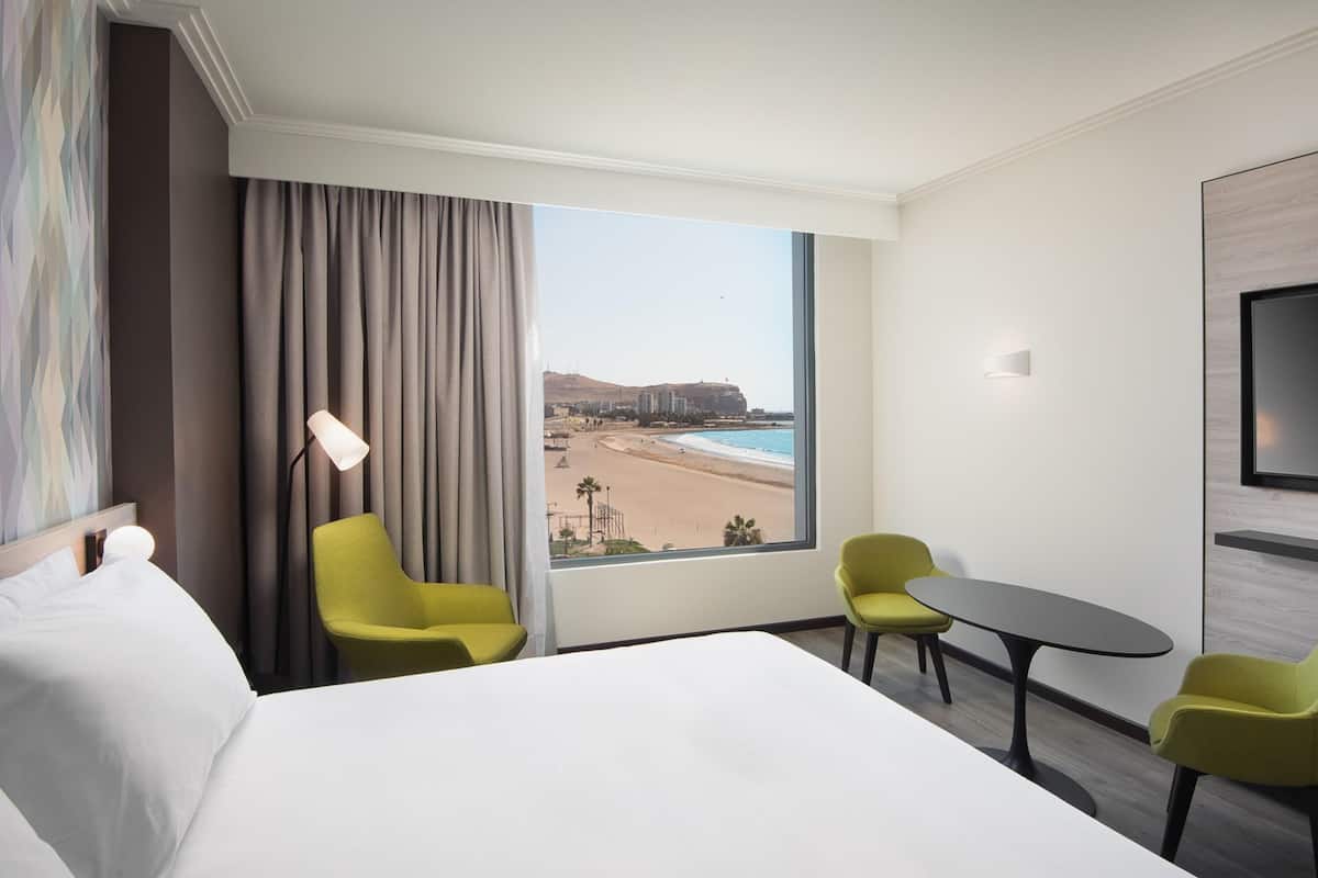 Ein Hotelzimmer mit einem Bett, zwei Stühlen, einem Tisch, einem Fernseher und einem großen Fenster mit Blick auf den Strand.
