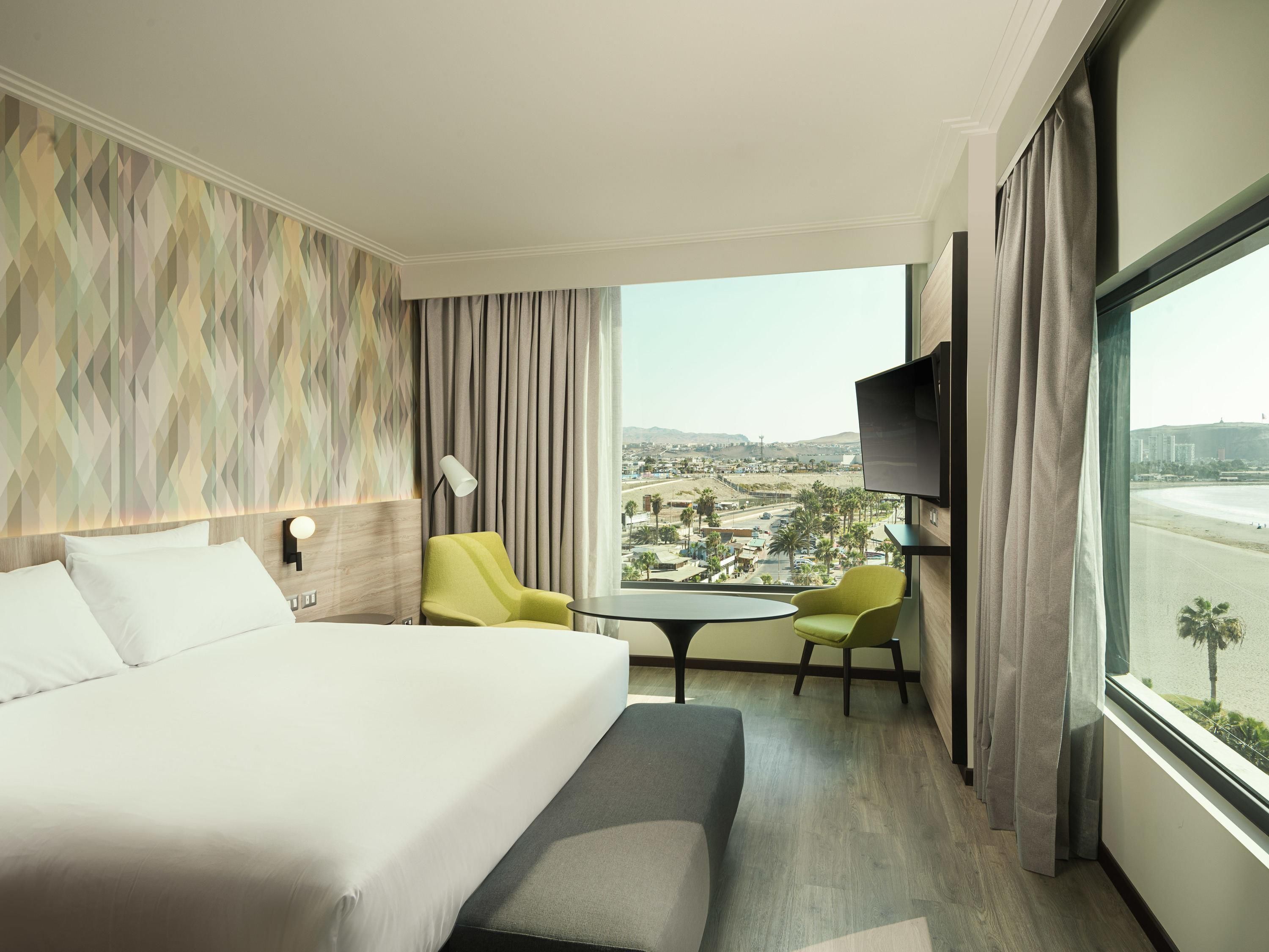 A hotel room with a bed, two chairs, a small table, a TV, and a large window with a view of a cityscape.