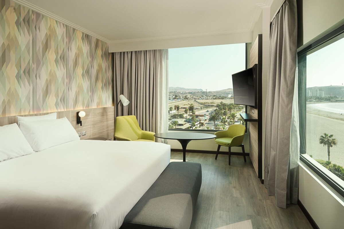 Habitación de hotel con cama, dos sillas, una mesita, televisor y ventana grande con vistas a la ciudad.