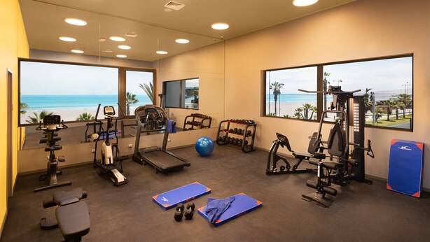 Un gimnasio con equipo de ejercicio, una colchoneta azul y vista a la playa.