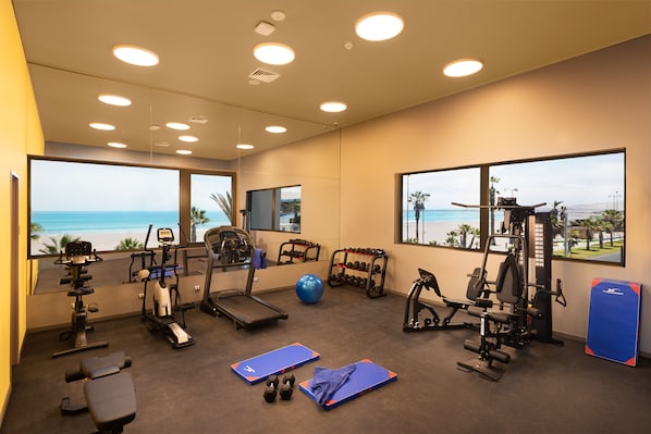 A gym with exercise equipment, a blue mat, and a view of the beach.