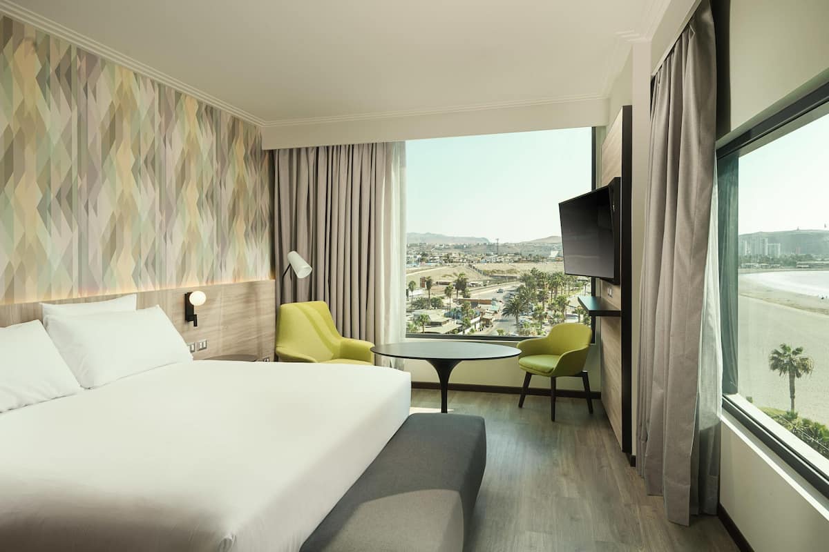 Ein Hotelzimmer mit einem Bett, zwei Sessel, einem kleinen Tisch, einem Fernseher und einem großen Fenster mit Blick auf die Stadt.