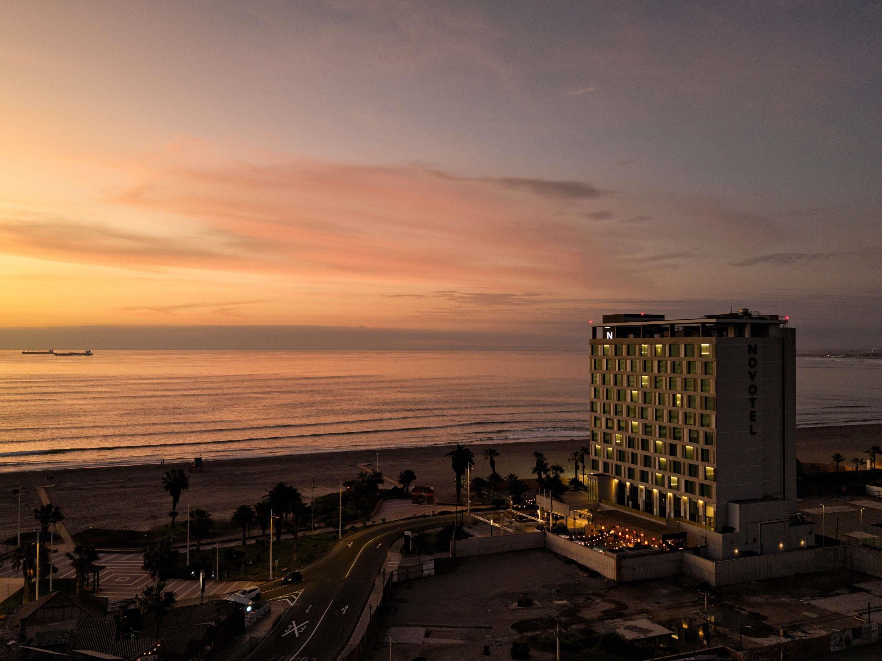A hotel with a view of the ocean at sunset, a parking area, and a pedestrian walkway.