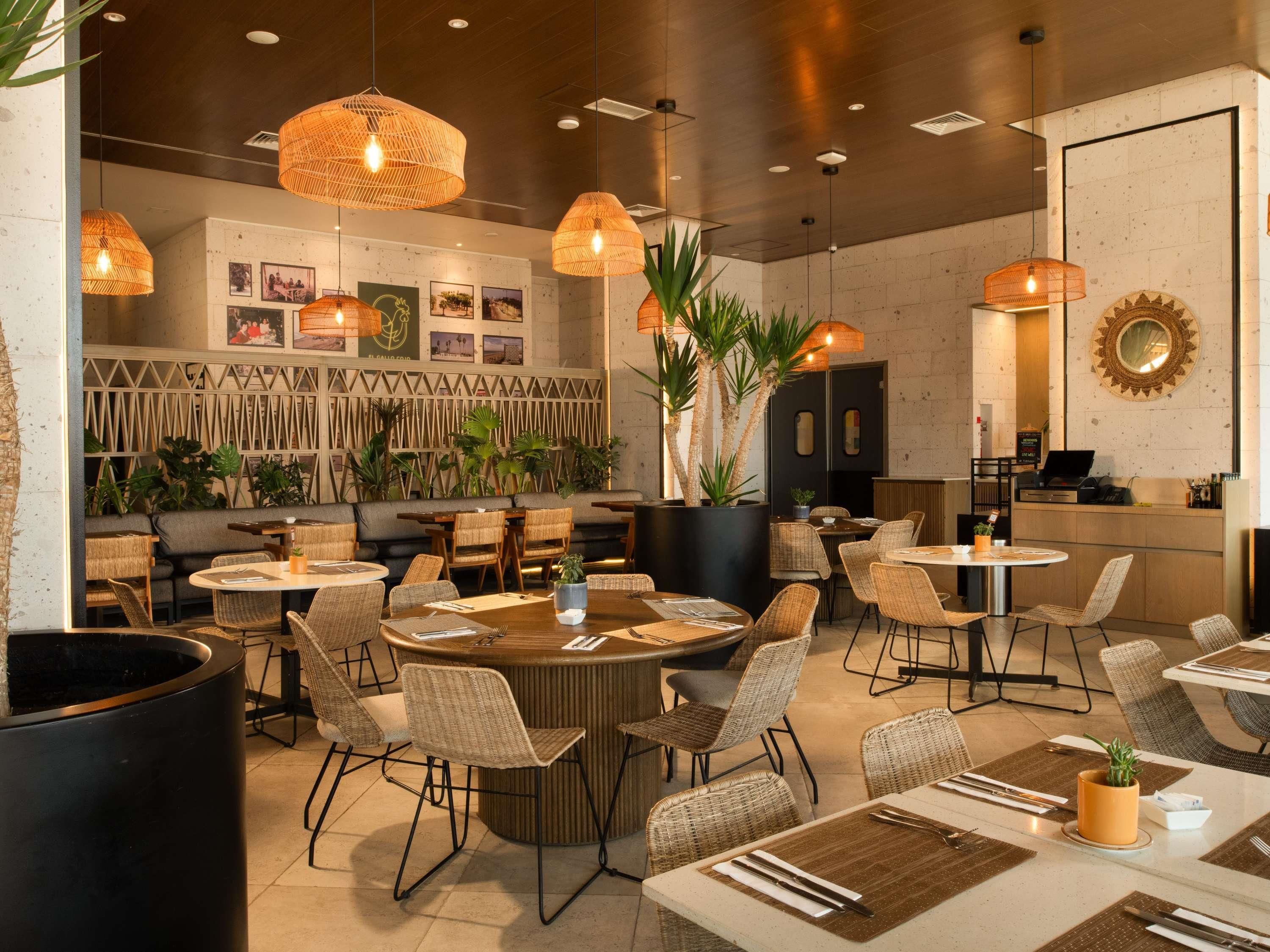 A restaurant interior with round tables, wicker chairs, hanging wicker lamps, and a wall with framed pictures.
