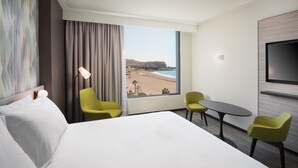 Habitación de hotel con una cama grande, dos sillas amarillas, una mesita, televisor y vista a la playa y al océano.