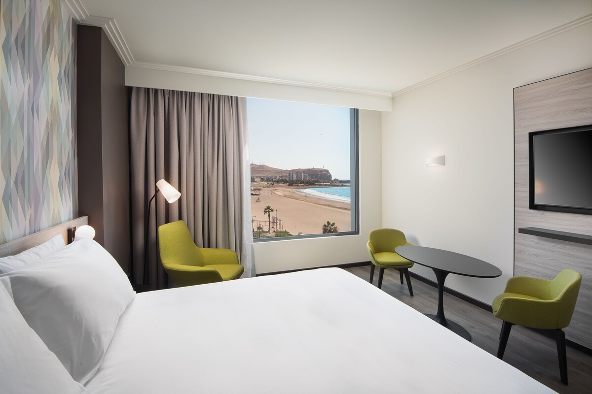 A hotel room with a large bed, two yellow chairs, a small table, a TV, and a view of a beach and ocean.
