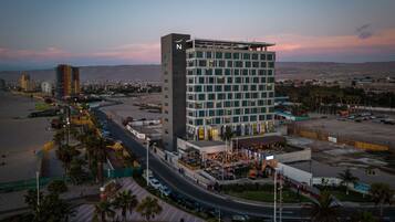 Ein hoch aufragendes Hotel mit modernem Design, das eine große 'N' an der Fassade zeigt, nahe einem Strand mit Parkplatz und Blick auf die Stadtlandschaft.