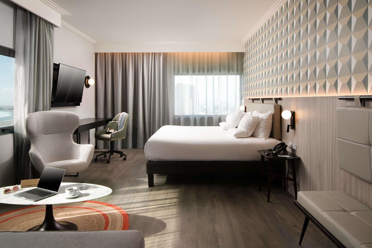 Habitación de hotel con cama, escritorio con una computadora portátil, silla, televisor y vista a la ciudad.