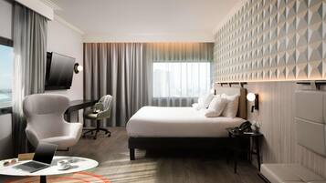 Habitación de hotel con cama, escritorio con una computadora portátil, silla, televisor y vista a la ciudad.