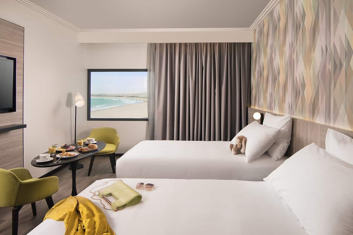 Ein Hotelzimmer mit zwei Betten, einem Schreibtisch mit Stuhl, einem Fernseher und Blick auf den Strand.