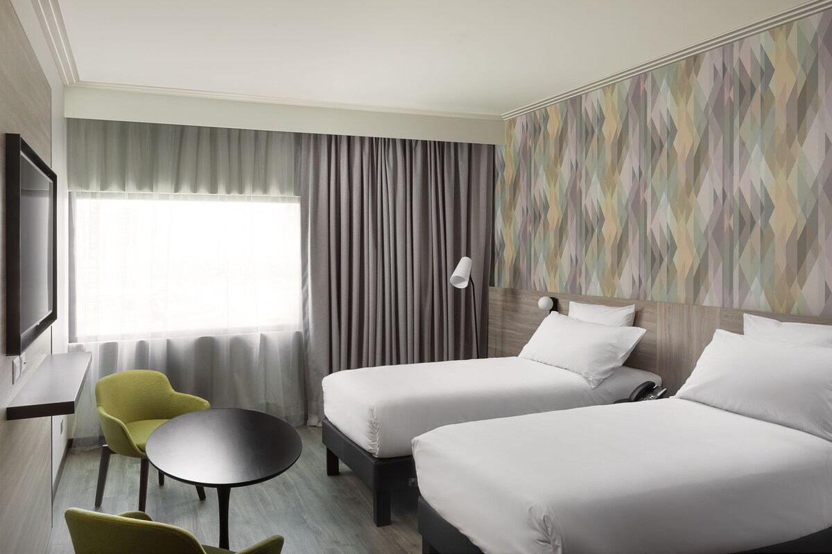 A hotel room with two beds, a small table, a chair, a TV, and a window with curtains.