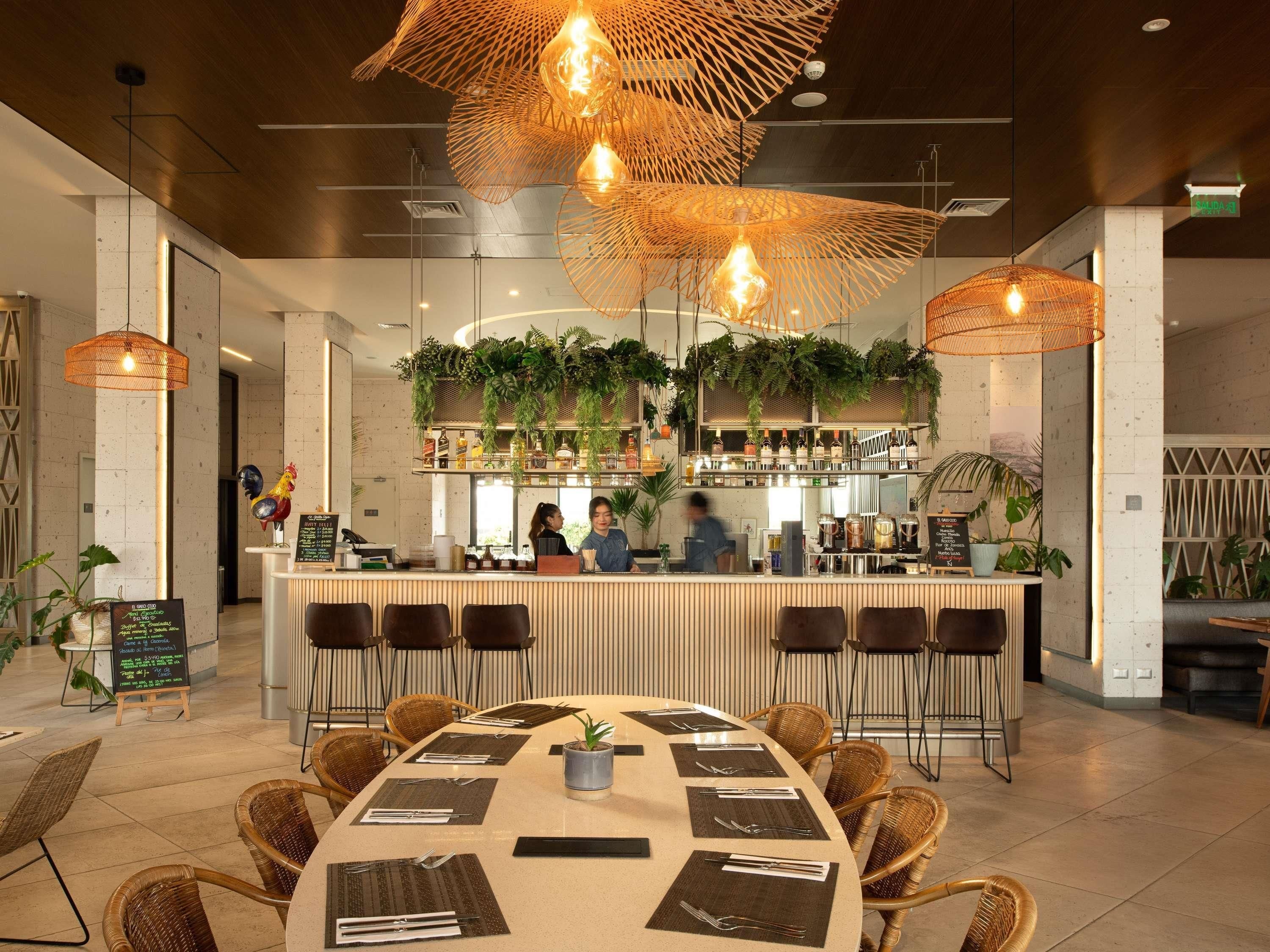 Ein modernes Restaurant mit einer zentralen Bar, Korbstühlen und hängenden Pflanzen.