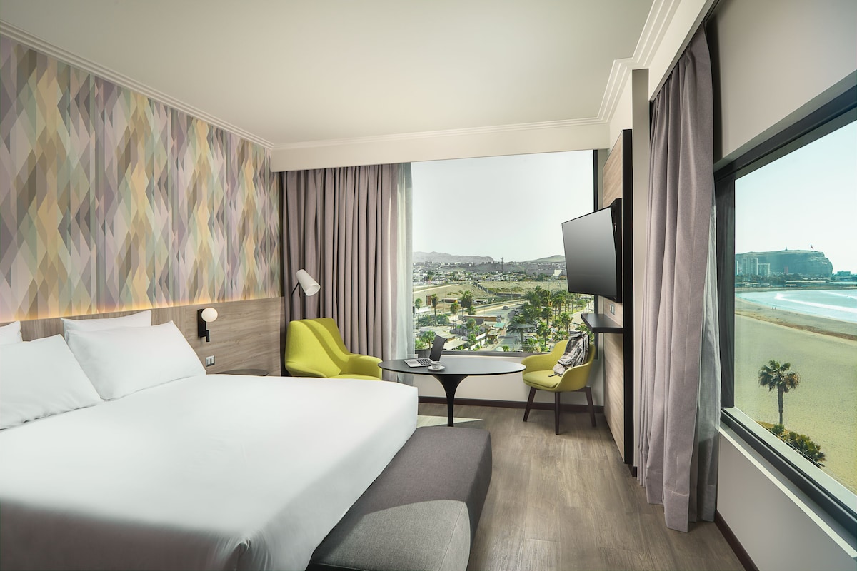 A hotel room with a bed, a desk, a chair, a TV, and a view of the beach.