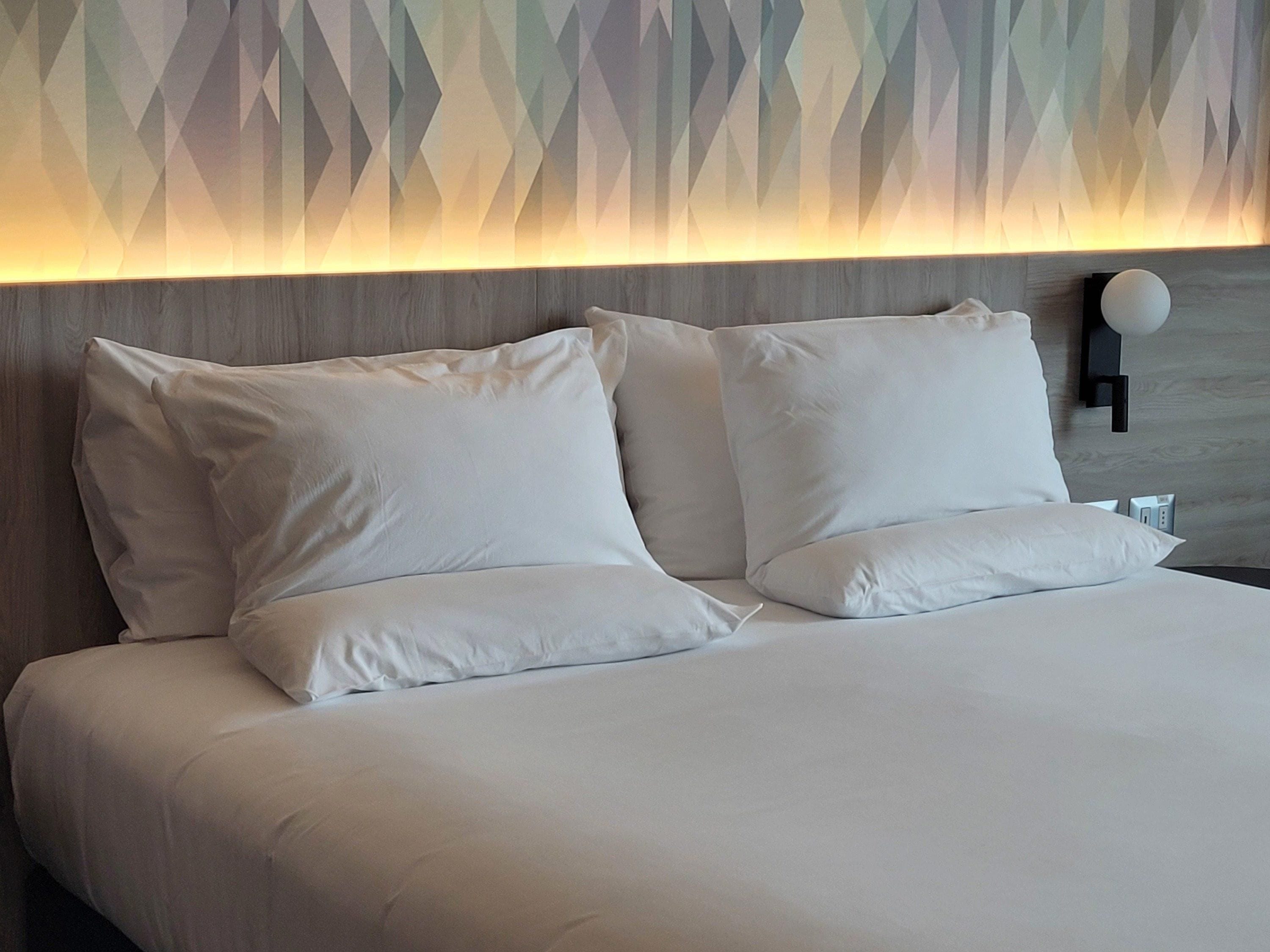 Ein ordentlich bezogenes Bett mit weißen Leinen und Kissen, dahinter eine Wand mit geometrischem Muster, und eine moderne Wandleuchte.