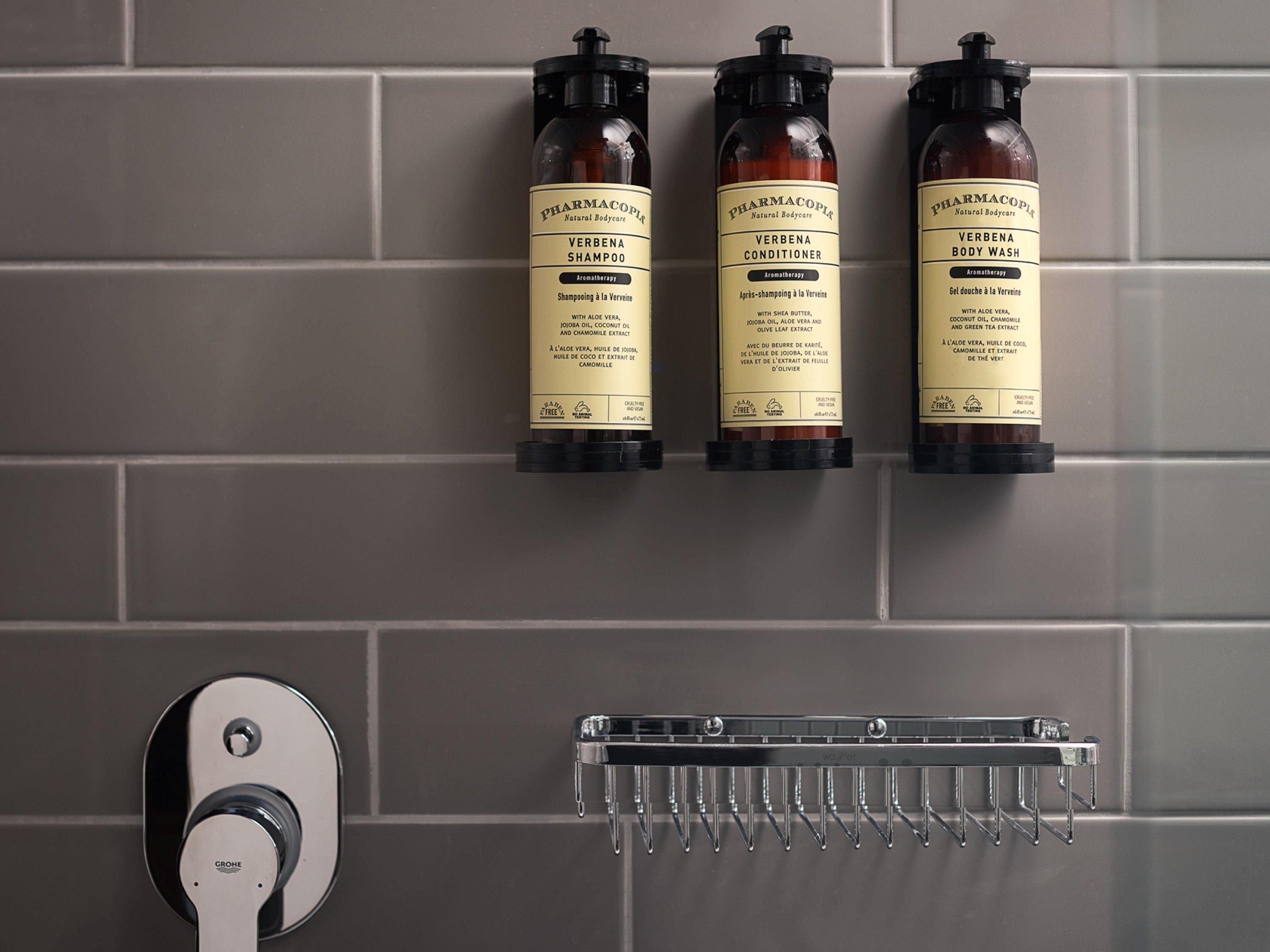 Three bottles of Verbena body wash and shampoo on a tiled wall.