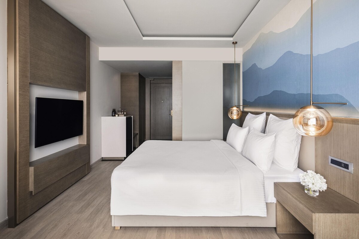 Une chambre d’hôtel moderne avec un grand lit, un téléviseur à écran plat fixé au mur, une table de chevet avec une lampe et un tableau représentant des montagnes accroché au mur.
