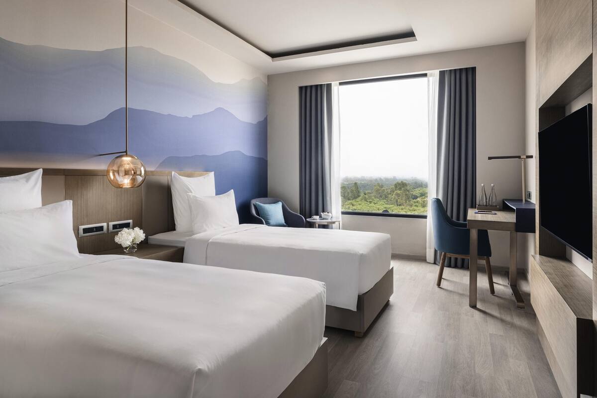 Habitación de hotel con dos camas, un escritorio, una silla, un televisor y un ventanal con vistas.