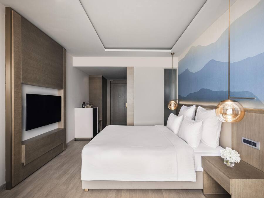 Een moderne hotelkamer met een groot bed, een flatscreen televisie aan de muur, een nachtkastje met een lamp en een berglandschap geschilderd op de muur.