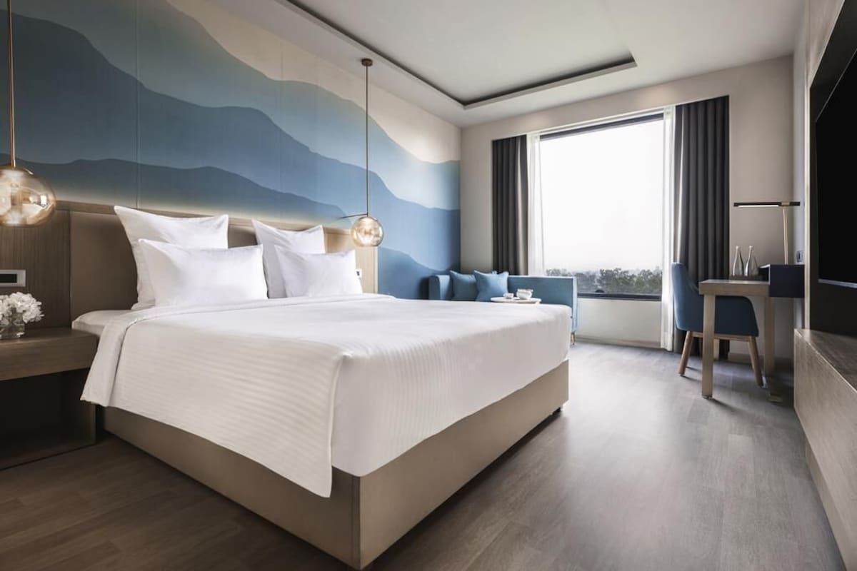 Ein modernes Hotelzimmer mit einem großen Bett, einem Schreibtisch, einem Sessel und Blick ins Freie, sowie einer dekorativen Wand.