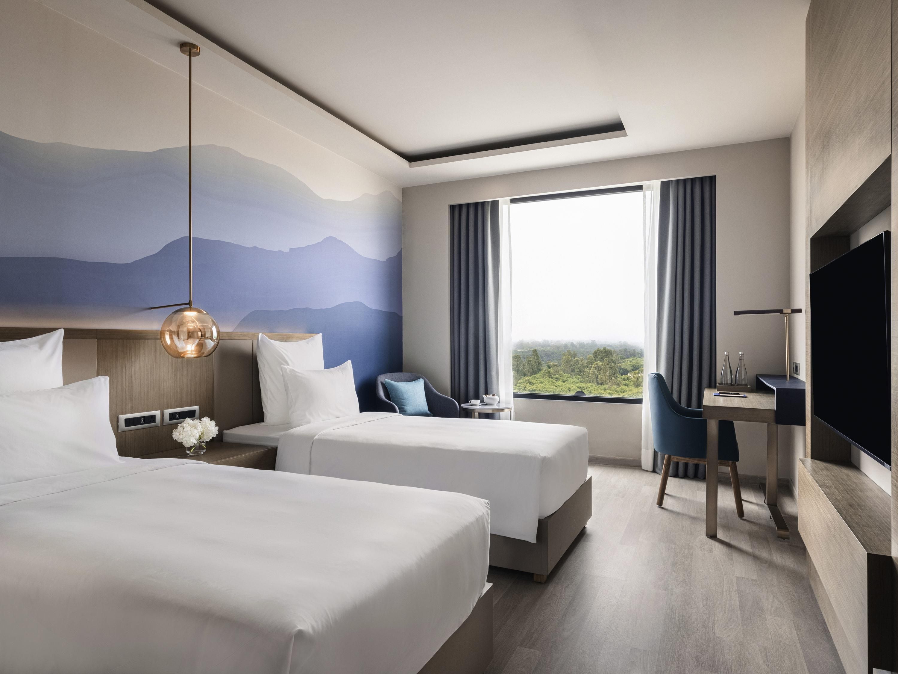 Habitación de hotel con dos camas, un escritorio, una silla, un televisor y un ventanal con vistas.
