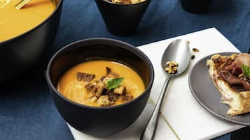 Een kom soep met croutons en een lepel, een bord met een stuk brood en vlees, en een kom ontbijtgranen met een lepel.