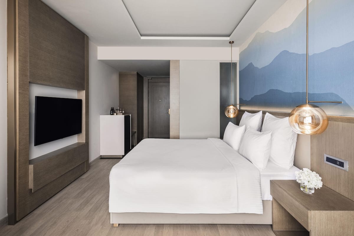 A modern hotel room with a large bed, a flat-screen TV mounted on the wall, a bedside table with a lamp, and a painting of mountains on the wall.