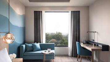 A modern hotel room with a bed, a sofa, a desk, and a view of the outdoors.