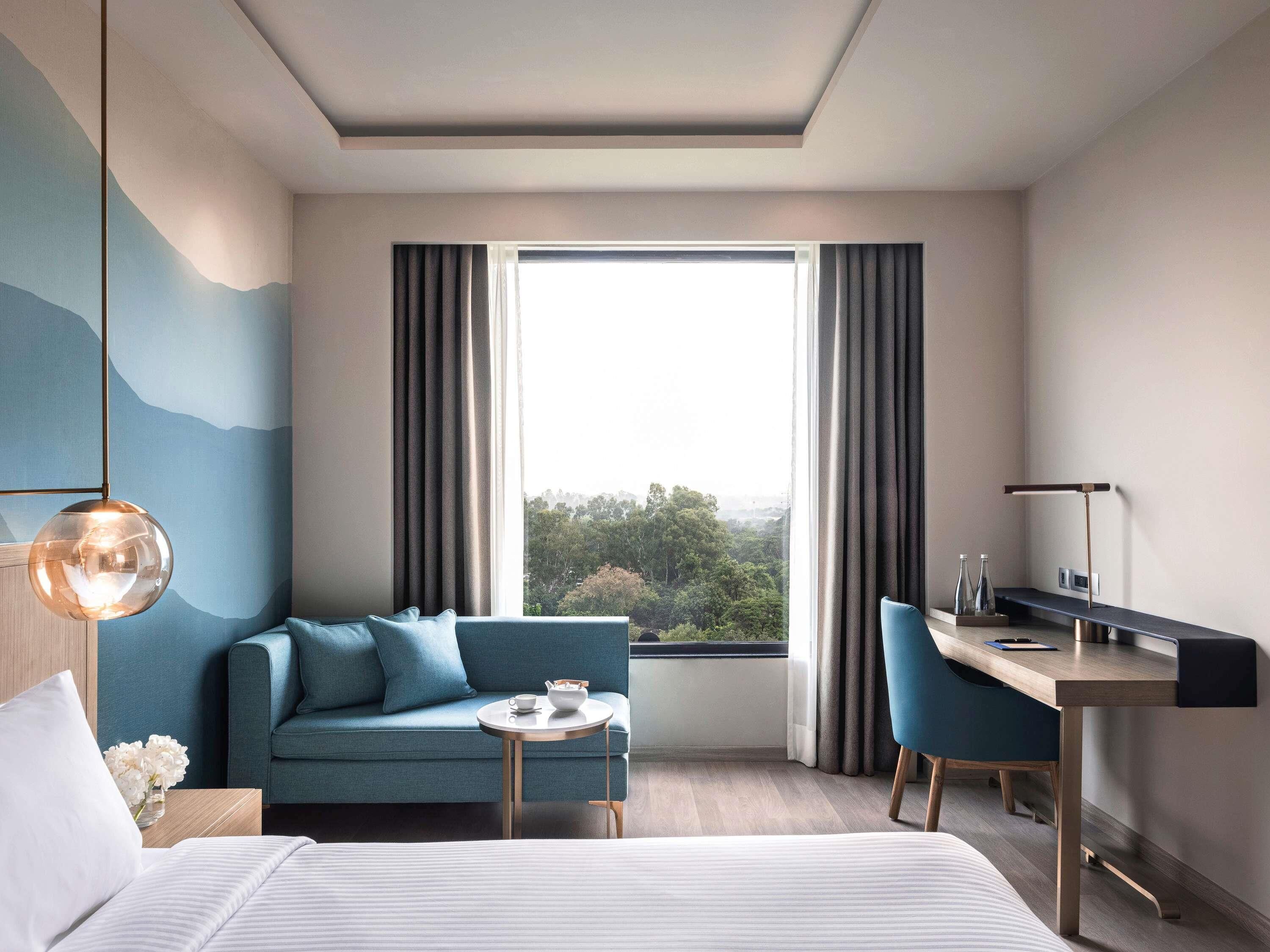 A modern hotel room with a bed, a sofa, a desk, and a view of the outdoors.