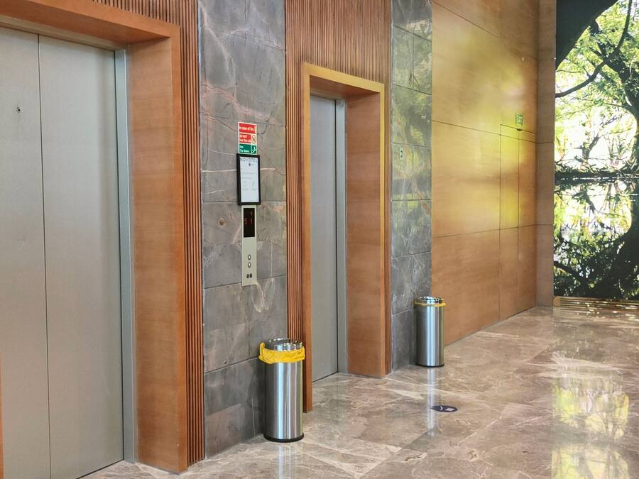 Two elevators with a digital display and a trash bin in a hallway with a polished floor and a mural of a tree.